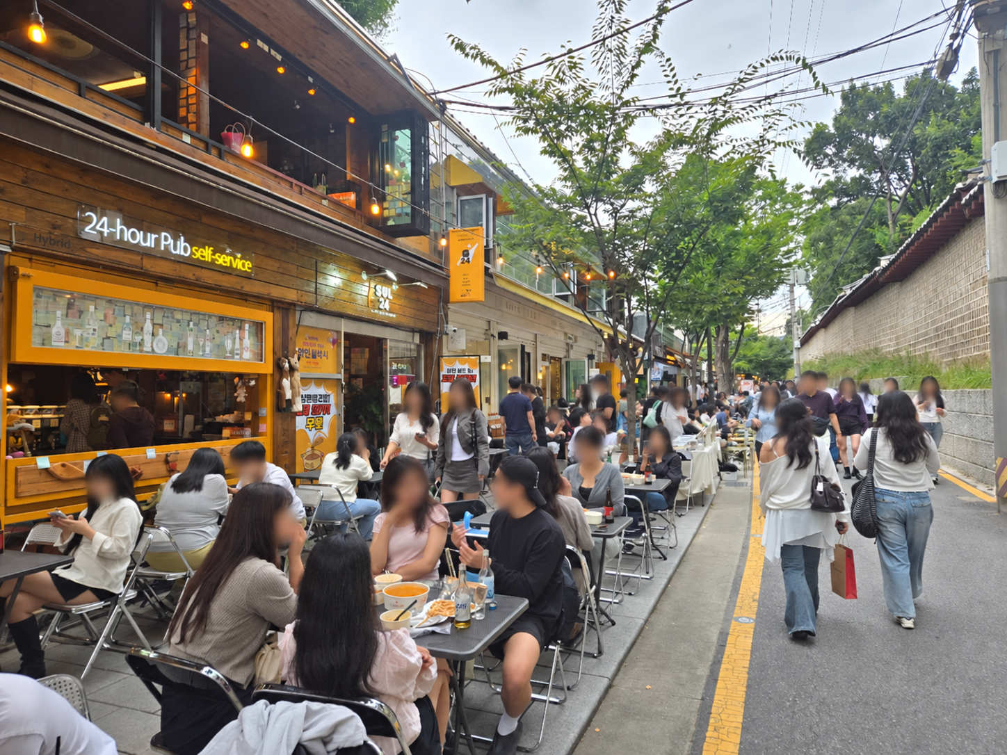 Busy outdoor seating and visitors on Seosulla-gil beside the Jongmyo wall; faces blurred for privacy